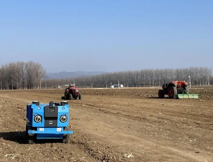 无驾驶舱拖拉机（红）正在作业，运输机器人（蓝）